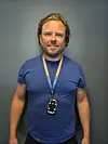 En mann i blå T-skjorte står foran en grå vegg med et ID-kort på snor rundt halsen.